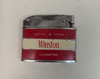 May include: Vintage Winston cigarette lighter with a red and white design. The lighter has a silver metal top and the word "Winston" is prominently displayed. The text "FILTER TIPPED" and "CIGARETTES" are also visible.