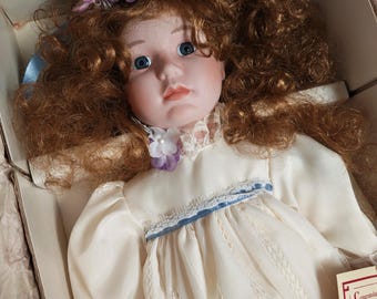 May include: A porcelain doll with auburn curly hair, blue eyes, and a fair complexion. The doll wears a cream-colored dress with lace and blue trim, and a floral hair accessory. The doll is in a cardboard box.