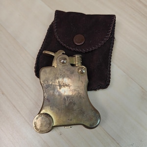 Puede incluir: Un encendedor antiguo de color latón con una forma única, acompañado de una funda de cuero marrón oscuro. El encendedor tiene un diseño distintivo con una palanca y una base circular. La funda tiene un cierre de botón.