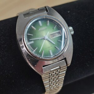 May include: A vintage Citizen automatic wristwatch with a silver-tone case and band. The watch face is a gradient green color with silver hands and markers. The watch displays the day and date. The watch is approximately 1.25 inches in diameter.