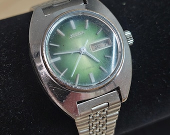 May include: A vintage Citizen automatic wristwatch with a silver-tone case and band. The watch face is a gradient green color with silver hands and markers. The watch displays the day and date. The watch is approximately 1.25 inches in diameter.