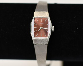 May include: A vintage Citizen watch with a rectangular, reddish-brown face and silver-toned band. The watch face features silver hands and markers. The watch has a textured finish on the band and case. The watch is displayed against a black background.