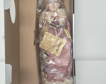 May include: A porcelain doll in a cardboard box, wrapped in plastic. The doll has blonde hair, blue eyes, and is wearing a pink dress with a lace collar. A tag is attached to the doll. The doll is labeled "Rapunzel".