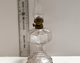 May include: A clear glass oil lamp with a brass burner and a glass chimney. The lamp has a hexagonal body and a decorative base. A ruler is placed next to the lamp.