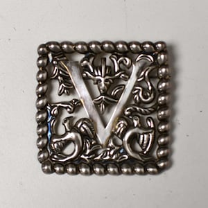 Peut inclure: Broche en métal argenté de forme carrée avec une bordure perlée. La broche présente la lettre "V" au centre, entourée de motifs floraux et animaliers stylisés et ornés. La broche est probablement vintage ou antique.