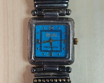 May include: A square-faced watch with a turquoise dial and silver-tone case. The watch face features the text "ECCUSSI" and "STERLING SILVER". The watch has a black leather band and gold-tone accents.
