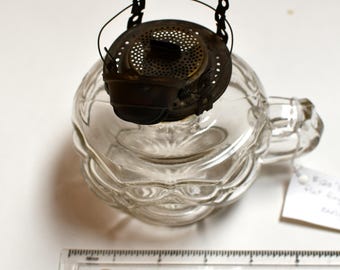 May include: Clear glass oil lamp with a decorative, round base and a metal top. The lamp has a handle and a metal wick holder. A ruler is in the foreground.