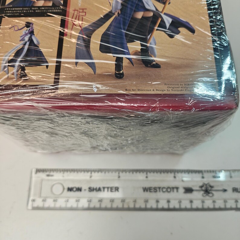 Puede incluir: Una figura de anime empaquetada, envuelta en pl&aacute;stico transparente. El arte de la caja presenta un personaje con un atuendo azul y blanco con tela larga y fluida. El texto en la caja incluye caracteres japoneses y las palabras "Presented by HIRO" y "Box Art Direction & Design by Noriyuki Zing". Una regla est&aacute; en primer plano.