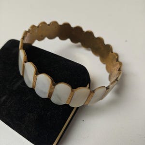 May include: A vintage brass bracelet with a scalloped edge design. The bracelet features alternating sections of brass and white, oval-shaped inlays. The bracelet is displayed on a black surface.