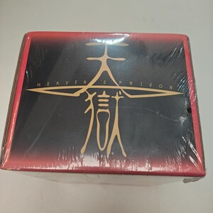 Puede incluir: Una caja rectangular envuelta en pl&aacute;stico transparente. La caja tiene un fondo negro con texto dorado que dice "HEAVEN'S PRISON" y un gr&aacute;fico dorado. La parte superior y los lados de la caja son rojos.