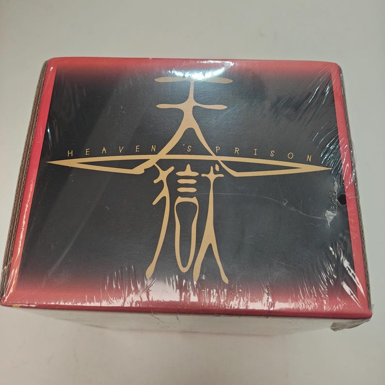 Puede incluir: Una caja rectangular envuelta en pl&aacute;stico transparente. La caja tiene un fondo negro con texto dorado que dice "HEAVEN'S PRISON" y un gr&aacute;fico dorado. La parte superior y los lados de la caja son rojos.