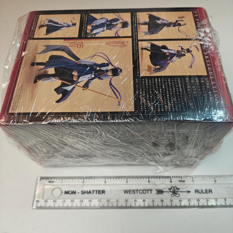 Puede incluir: Una caja rectangular envuelta en pl&aacute;stico transparente, con ilustraciones de un personaje con un atuendo fluido con una espada. La caja tiene texto japon&eacute;s y las palabras "ARTICULATION" y "23 PON". Una regla Westcott est&aacute; en primer plano.