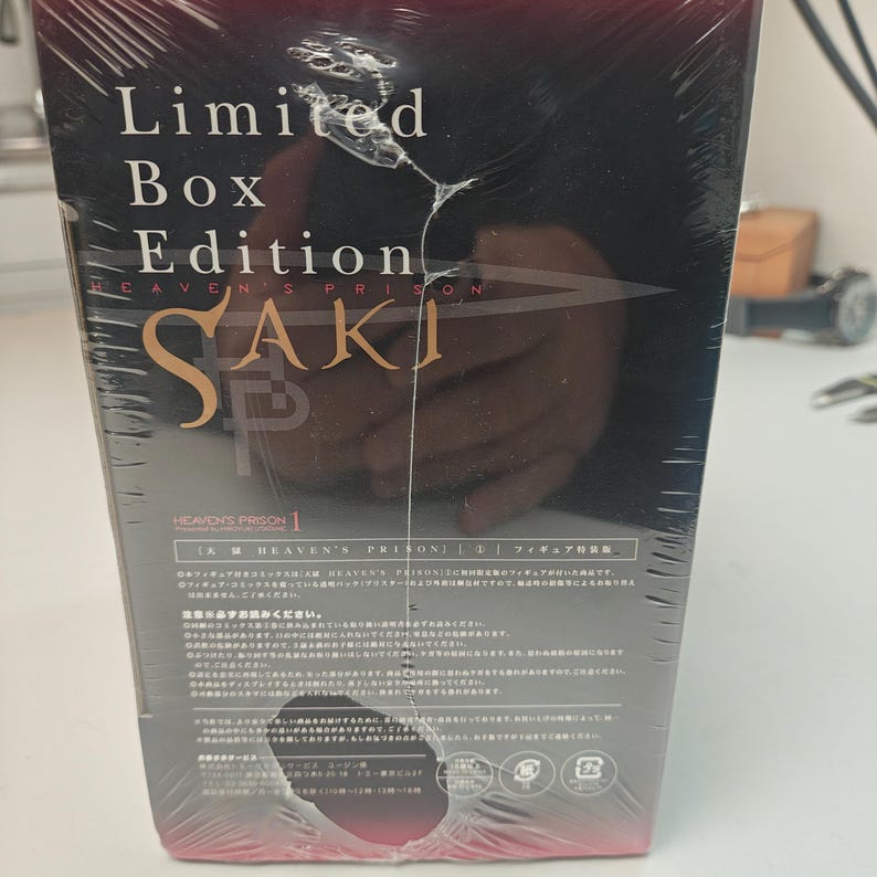 Puede incluir: Una edici&oacute;n limitada y sellada de "Heaven's Prison Saki" con texto en japon&eacute;s. La caja est&aacute; envuelta en pl&aacute;stico transparente y presenta el t&iacute;tulo en letras doradas. El fondo es oscuro con una imagen borrosa.