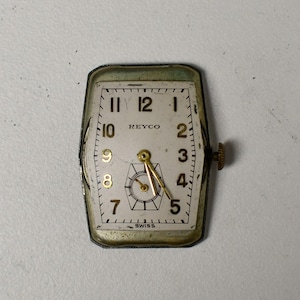 May include: A vintage rectangular watch face with a white dial, black numerals, and gold hands. The watch face is marked "REYCO" and "SWISS". The metal casing has a tarnished, aged appearance, and a small crown is visible on the side.