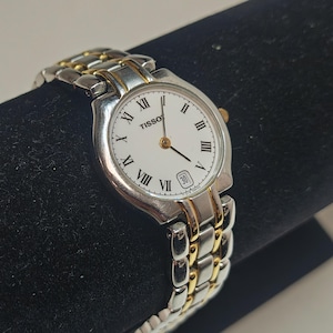 May include: A Tissot wristwatch with a white face, gold hands, and Roman numeral hour markers. The watch has a silver and gold-tone metal band and a date display at the 3 o'clock position. The watch is displayed on a black surface.