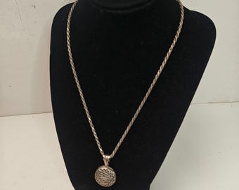 May include: A silver-toned rope chain necklace featuring a circular pendant with an intricate, openwork filigree design. The pendant has a textured, metallic finish and is suspended from a twisted metal chain that secures around the neck.