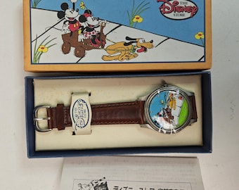 May include: A Disney Store wristwatch with a brown leather strap and a round silver-tone case. The watch face features Mickey Mouse, Minnie Mouse, and Pluto. It is presented in a rectangular blue and tan box with a matching character illustration.