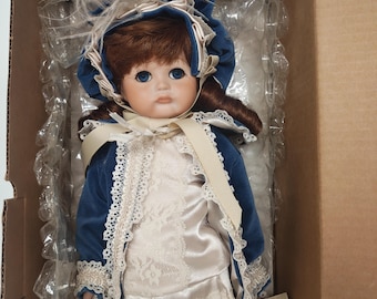 May include: A porcelain doll with blue eyes and auburn hair, wearing a blue velvet coat with lace trim and a matching bonnet adorned with feathers. The doll is dressed in a cream-colored dress and is packaged in a cardboard box.