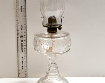 May include: A clear glass oil lamp with a decorative etched design around the reservoir. The lamp has a clear glass chimney and a metal burner assembly. A ruler is placed next to the lamp. The lamp is approximately 10 inches tall.