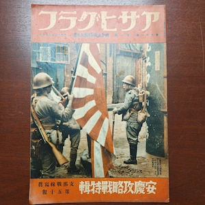 May include: Vintage Japanese magazine cover featuring soldiers and a rising sun flag. The cover is predominantly orange and red, with Japanese text at the top and bottom. Soldiers in uniform are depicted in a street scene, holding rifles and the flag.
