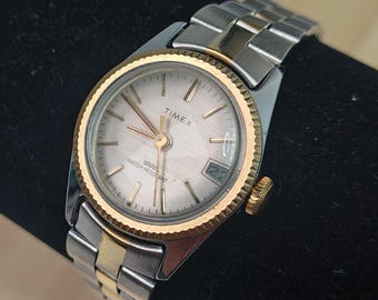 May include: A vintage Timex watch with a silver dial, gold-tone bezel, and a two-tone silver and gold metal band. The watch face displays the brand name, the date, and the words "Water Resistant". The watch is displayed on a black surface.