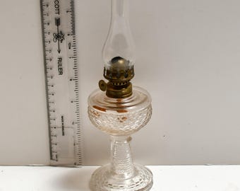 May include: A small, clear glass oil lamp with a textured base and a brass-colored burner. The lamp has a glass chimney and a small, round fuel reservoir. A ruler is placed next to the lamp.