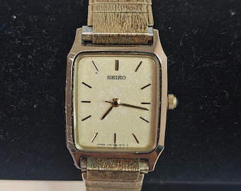 May include: A vintage Seiko watch with a gold-toned rectangular face and matching band. The watch face is a light champagne color with black hour markers and hands. The watch band has a textured, woven appearance. The watch is set against a dark background.