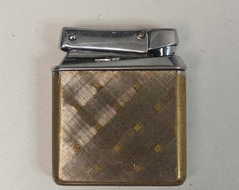 May include: Vintage gold-toned metal lighter with a silver-colored top. The lighter has a textured, cross-hatched pattern on the front and sides, with small square accents. The top is a shiny silver color.