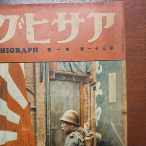 May include: Vintage Japanese magazine cover with Japanese text in white and orange. The cover features a soldier in a helmet and military gear, with a Japanese flag in the background. The magazine is titled "HIGRAPH".