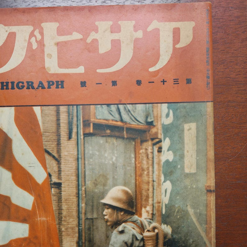 May include: Vintage Japanese magazine cover with Japanese text in white and orange. The cover features a soldier in a helmet and military gear, with a Japanese flag in the background. The magazine is titled "HIGRAPH".
