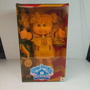 Può includere: Bambola Cabbage Patch Kids OlympiKids Special Edition nella sua scatola originale. La bambola indossa un completo da tennis giallo con dettagli rossi e blu e il logo olimpico USA. La scatola presenta il logo Cabbage Patch Kids e il testo "Tennis Olympics".