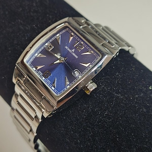 May include: A silver Jacques Lemans wristwatch with a rectangular face and a blue dial. The watch has silver hands and markers, a date display, and a stainless steel strap. The watch is displayed on a black surface.