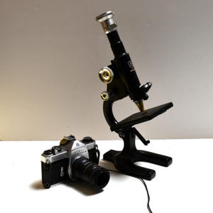 May include: A black microscope with silver accents stands next to a silver and black Pentax camera with a black lens. The microscope has a silver eyepiece and a black base. The camera has a black lens and a silver body.