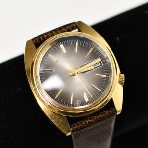 Puede incluir: Un reloj de pulsera dorado con una correa de cuero marrón. La esfera del reloj es gris con agujas e indicadores dorados. El reloj tiene el texto "SICURA 17 JEWELS AUTOMATIC SWISS MADE" en la esfera y una pantalla de fecha.