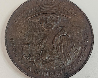 May include: A bronze-colored Coca-Cola Company round metal sign. The sign features a woman in a hat holding a glass, with the text "Coca-Cola" at the top and "Drink at Fountains 5¢" and "Coca-Cola Company Atlanta GA" around the edges.