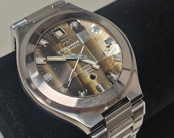 May include: A silver-toned Technos Automatic wristwatch with a metallic brown dial featuring a date window at the twelve o'clock position. The watch has a stainless steel link bracelet, silver hands, and rectangular hour markers on the face.