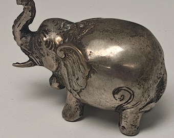May include: A small, silver-colored elephant figurine with its trunk raised. The figurine has detailed features, including ears, tusks, and wrinkles. The elephant is standing on four legs and has a curled tail.