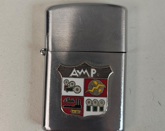 May include: A silver-colored metal lighter with a decorative emblem. The emblem features the letters "AMP" and various colorful illustrations, including film reels and industrial elements. The lighter has a hinged lid and a small metal hinge.