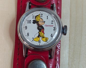 May include: A vintage Ingersoll wristwatch with a red leather band. The watch face features Mickey Mouse with black, red, yellow, and white colors. The watch has a silver-tone metal case and a black crown. The band is secured with silver-tone metal rivets.