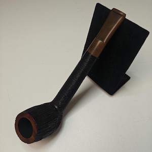 以下が含まれることがあります： 暗く質感のあるボウルと、長く黒く巻かれたステムを備えた手作りのタバコパイプ。 マウスピースは明るい茶色です。 パイプは白い背景に対して黒いスタンドに表示されています。