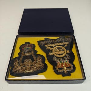 May include: Two embroidered military patches in a navy blue box. The patches are black with gold and colorful thread details. One patch features a crown and the other has a cannon design with the text "J-G-HOOK 1976".
