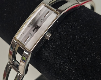 May include: A silver-tone Citizen XC wristwatch with a rectangular face, a light-colored dial with vertical brushed texture, and a slim, open-link metal bracelet. The watch face features the text XC and CITIZEN with thin black hands.