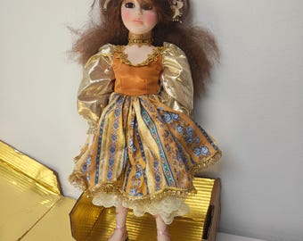 May include: A porcelain doll with brown hair and a floral crown. The doll wears a gold and orange dress with a patterned skirt, gold sleeves, and a gold choker. The doll is standing on a gold surface.