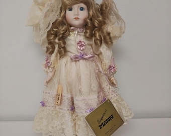 May include: A porcelain doll named "Jenny" with blonde curly hair, blue eyes, and a cream-colored lace dress with lavender accents. The doll has a tag that says "Seymour Mann". The doll is wearing cream-colored shoes with pink roses.