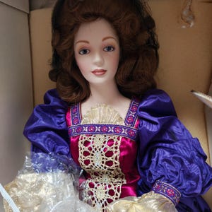 Może przedstawiać: Porcelanowa lalka z brązowymi włosami i niebieskimi oczami, ubrana w fioletowo-magentową suknię ze złotymi akcentami. Suknia ma koronkowe body i bufiaste rękawy. Lalka jest w pudełku.