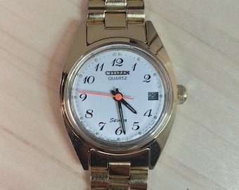 May include: A gold-toned Citizen Quartz watch with a white face, black numerals, and a date display at the 3 o'clock position. The watch has a gold-toned band and hands, with an orange second hand. The watch is on a light brown surface.