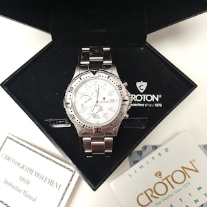 May include: A silver Croton chronograph watch with a white face, presented in a black box. The watch has multiple dials and a stainless steel strap. The open box displays the watch and paperwork. The text "CROTON" is visible on the watch and box.