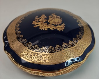 May include: A navy blue ceramic trinket box with a gold rim and decorative accents. The lid features a gold illustration of two figures in a garden setting. The box has a scalloped edge and is likely vintage.