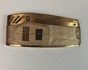 May include: Vintage gold-toned metal lighter with a textured, off-white leather wrap. The lighter features a rectangular shape with a hinged top and decorative cutouts. Two rectangular insets are visible on the leather.