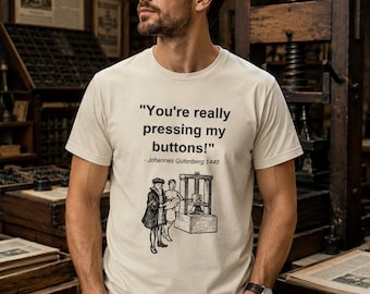 May include: A cream-colored short-sleeve t-shirt featuring black text that reads "You're really pressing my buttons!" followed by "- Johannes Gutenberg 1440" and a black-and-white illustration of two figures standing next to a printing press.